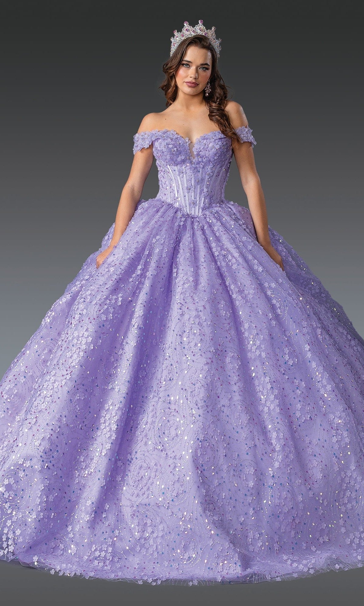 A woman stands gracefully against a plain gray background, wearing the Quinceanera Dress 1901 by Dancing Queen in sparkling lilac with off-the-shoulder sleeves and a jeweled crown.